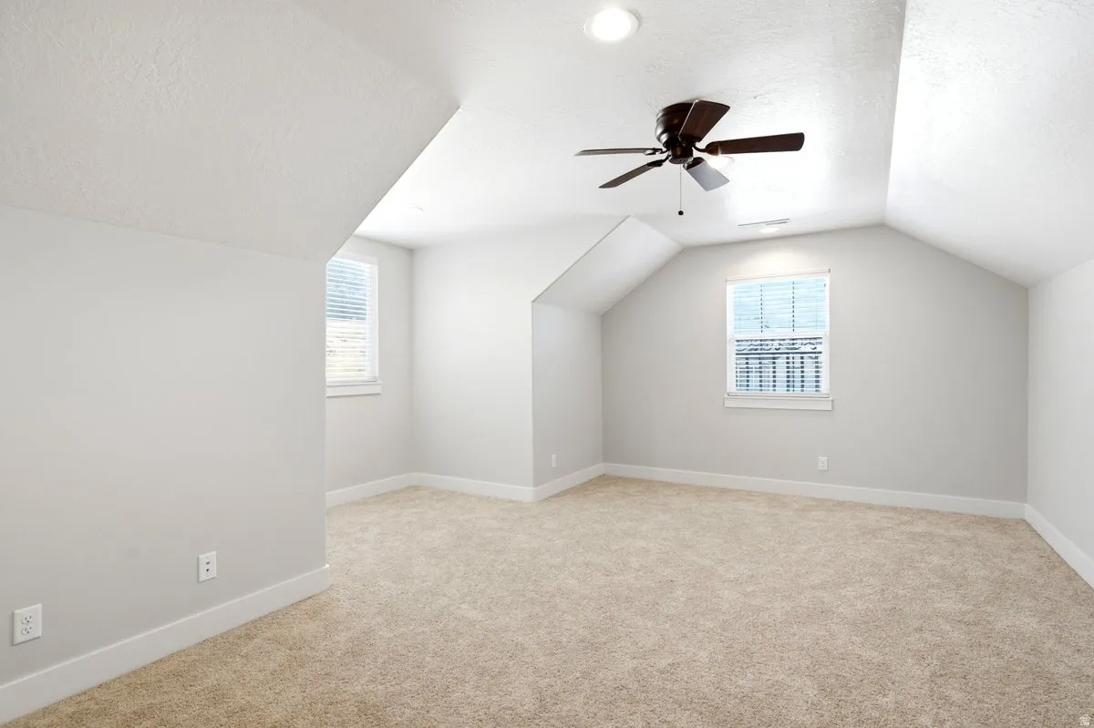 Spacious room featuring vaulted ceilings, neutral-toned carpeting, two windows with blinds, a ceiling fan with integrated lighting, and recessed ceiling lights