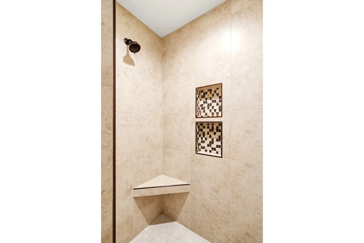 Tiled shower enclosure featuring a bronze showerhead, two recessed shelving niches with decorative mosaic tile accents, and a corner bench seat
