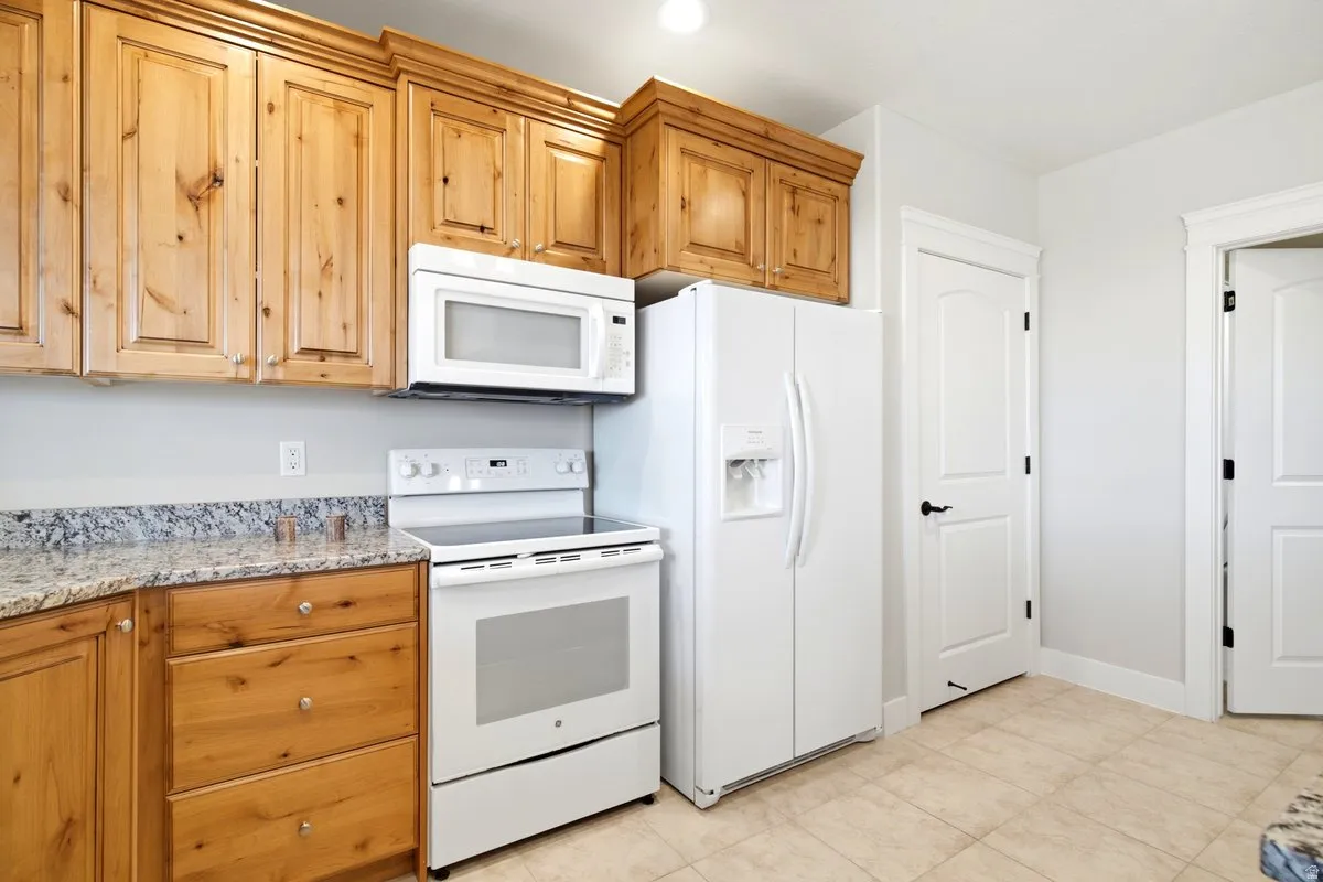 Kitchen featuring wood cabinetry, light-toned granite-style countertops, white appliances including a range and microwave, and tile flooring