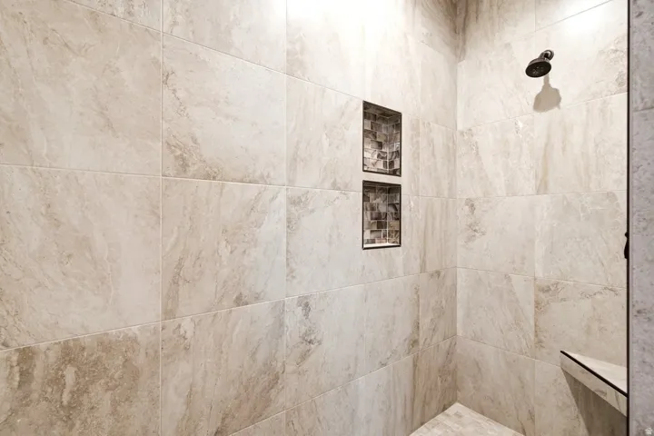 Walk-in shower with large-format beige tiles, two recessed mosaic tile niches, and a corner bench