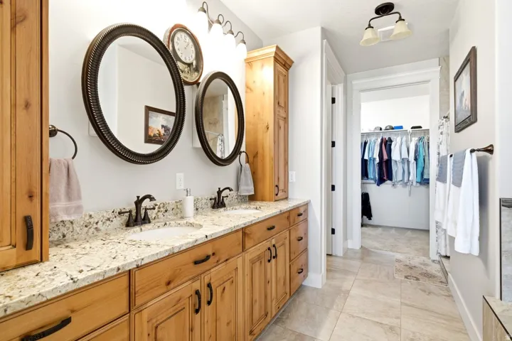 Double vanity with light-colored speckled countertops, twin oval mirrors with dark frames, wood-finish cabinetry, and a walk-in closet beyond