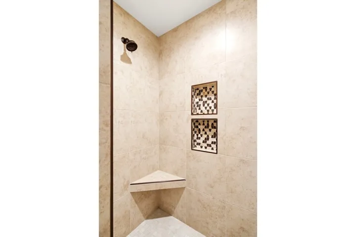 Tiled shower enclosure featuring a bronze showerhead, two recessed shelving niches with decorative mosaic tile accents, and a corner bench seat