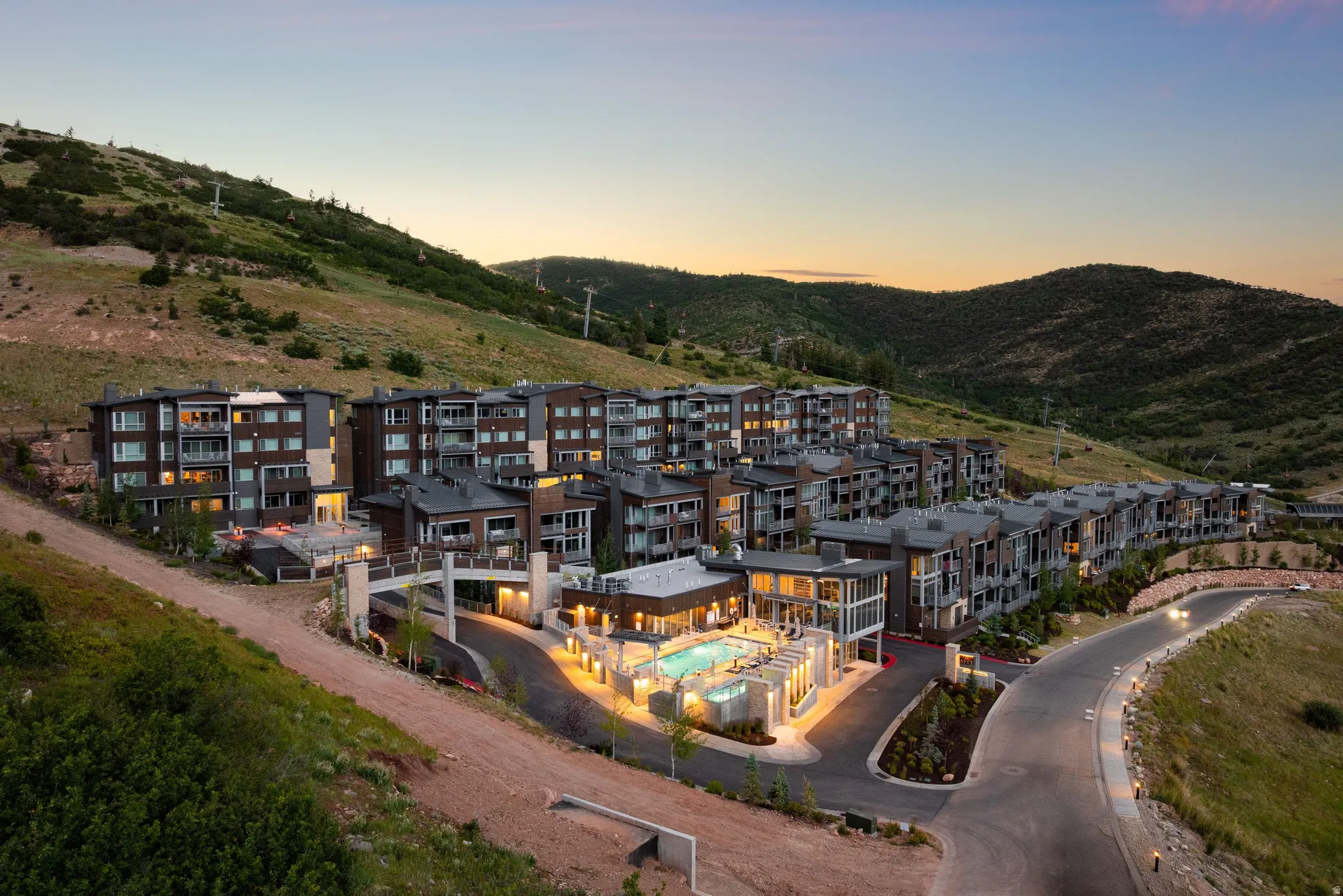 Modern hillside condominium complex featuring multiple buildings with private balconies, a central amenity building with an outdoor pool and hot tub, and paved access roads