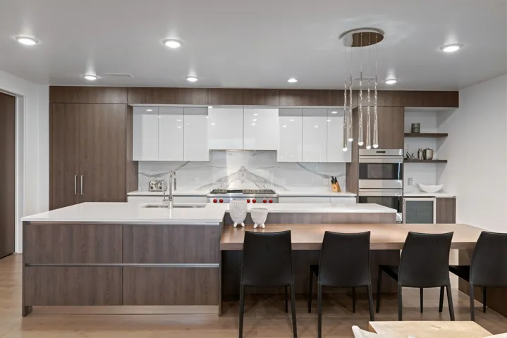Contemporary kitchen featuring wood-finish cabinetry, white high-gloss upper cabinets, a stone countertop island, and a distinctive linear chandelier