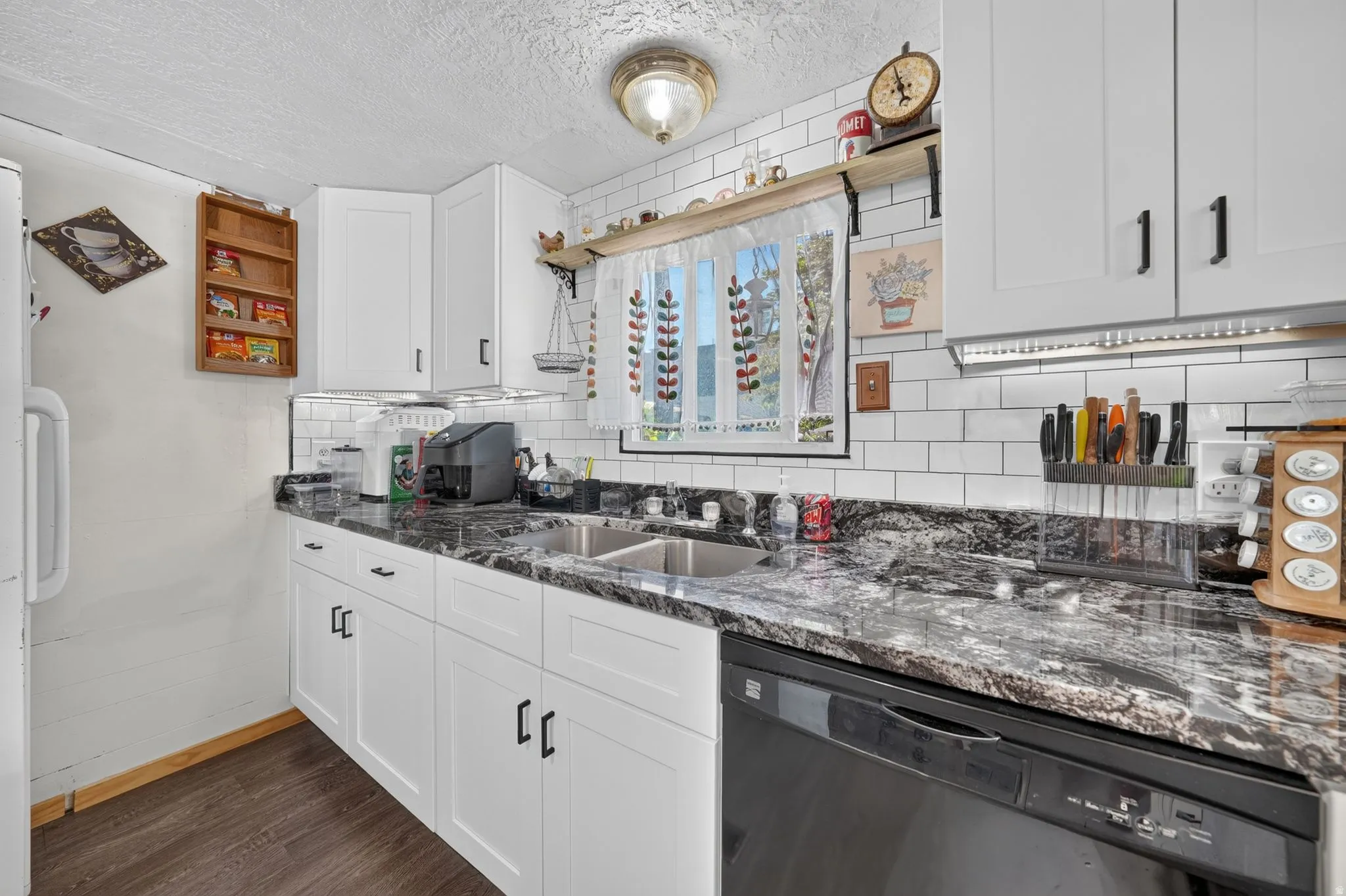 Kitchen featuring white shaker-style cabinetry with matte black hardware, dual-basin stainless steel sink, black and white patterned countertops, subway tile backsplash, and dark wood-finish flooring