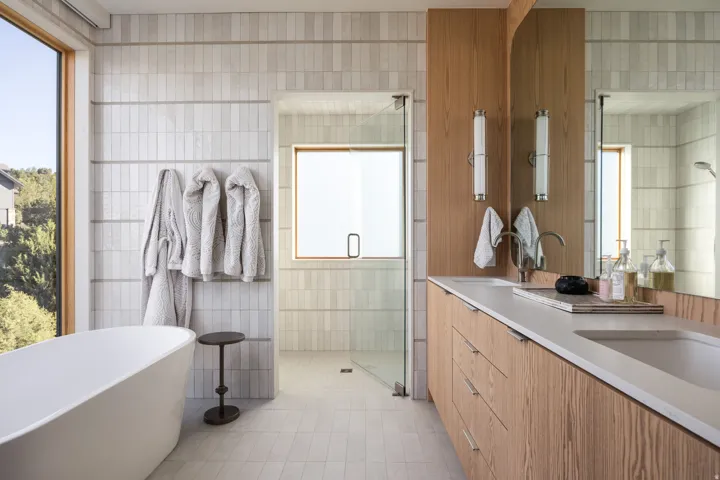 Contemporary bathroom featuring a freestanding soaking tub, wood-finish vanity with dual integrated sinks, glass-enclosed shower, and large format window