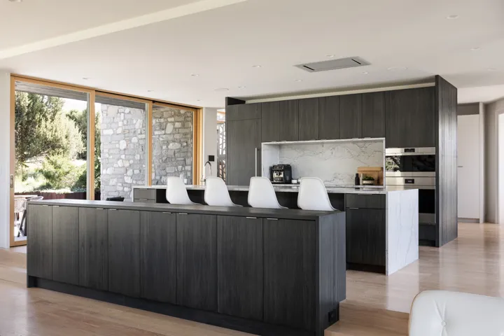 Modern kitchen featuring extensive dark wood-finish cabinetry, a large waterfall-edge island with a stone countertop, integrated stainless steel appliances, and a full-height stone backsplash