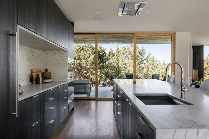 Gourmet kitchen featuring dark wood-finish cabinetry, light-toned stone countertops, integrated stainless steel appliances, a large center island with an undermount sink, and expansive glass sliding doors