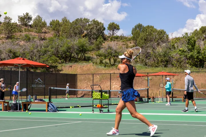 Tennis court featuring a green playing surface, black perimeter fencing, and red sun umbrellas