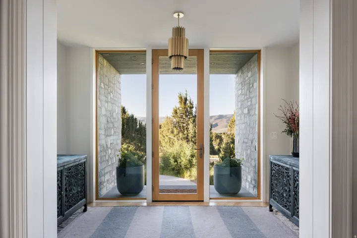 Entryway featuring a natural wood frame glass door, flanking floor-to-ceiling side windows with stone masonry surrounds, a modern brass chandelier, and a striped area rug
