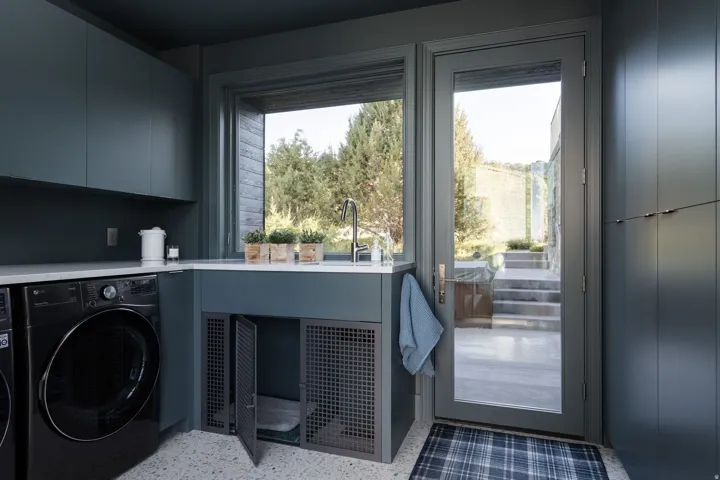 Contemporary laundry area featuring dark cabinetry, a built-in sink with a gooseneck faucet, light-toned countertops, and a full-light glass door leading to exterior stone steps