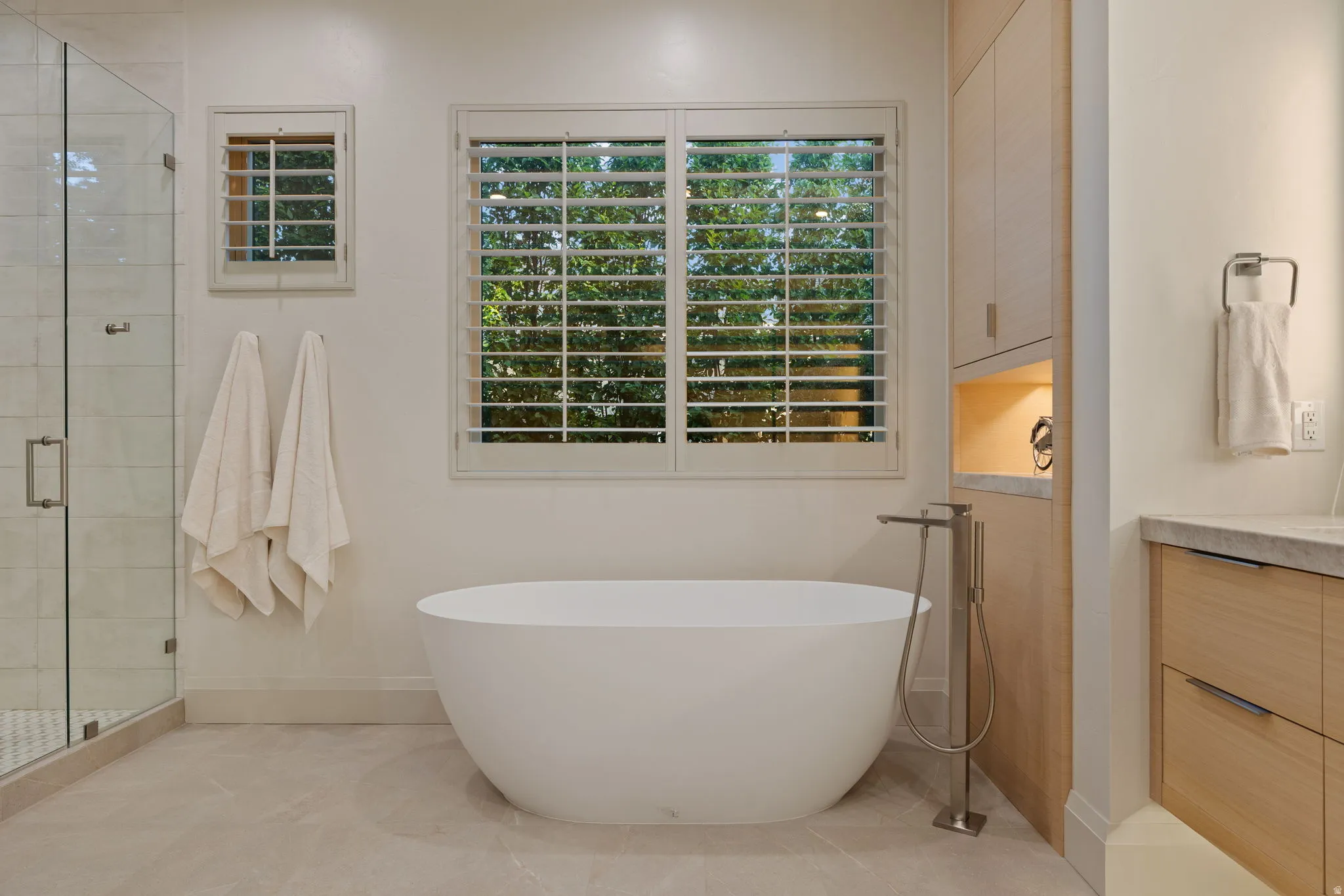 Freestanding soaking tub with floor-mounted faucet, plantation shutters, and a glass-enclosed shower with a tiled interior