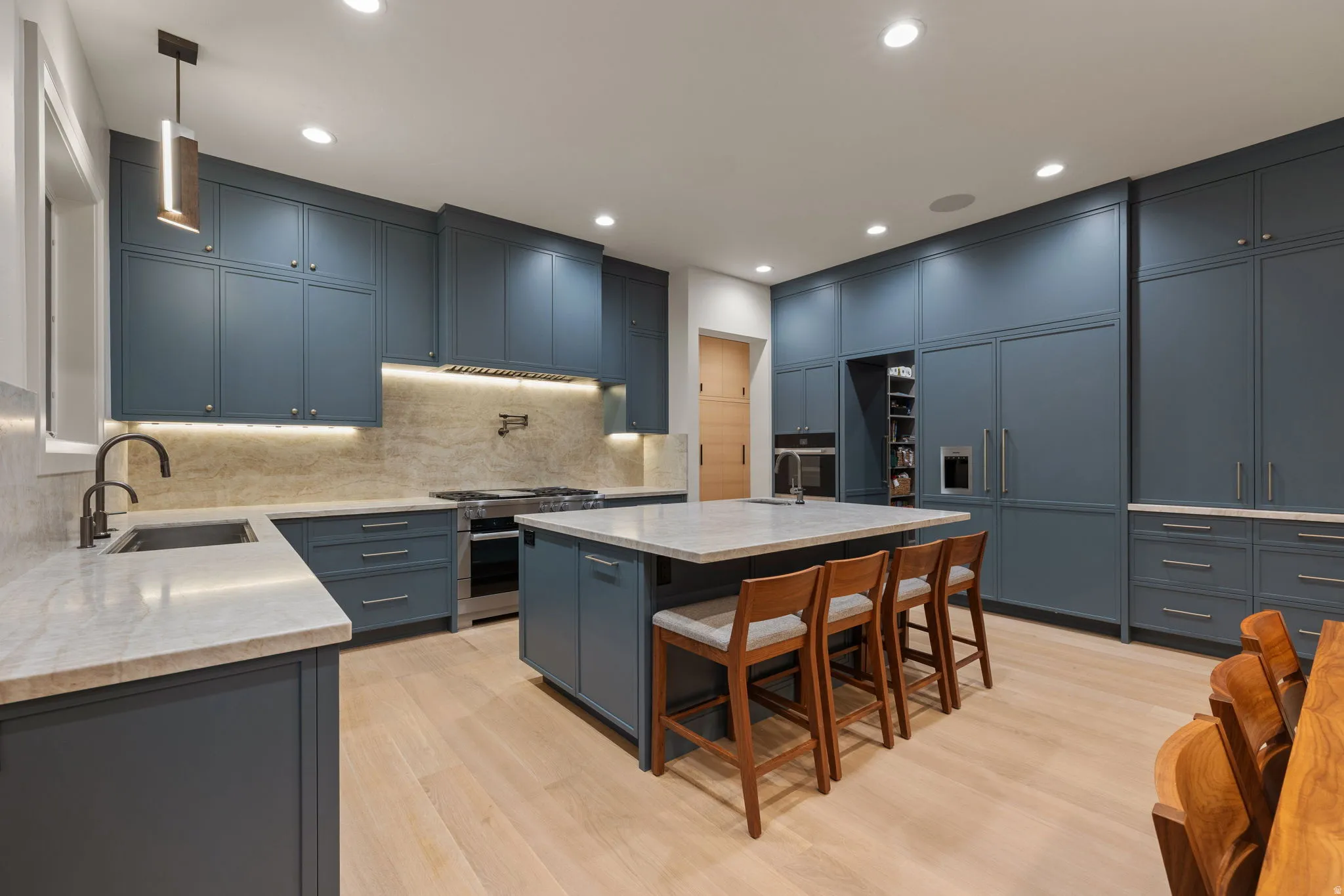 Spacious kitchen featuring extensive blue cabinetry, a large center island with seating, integrated stainless steel appliances, light-toned wood-finish flooring, and light-colored stone countertops
