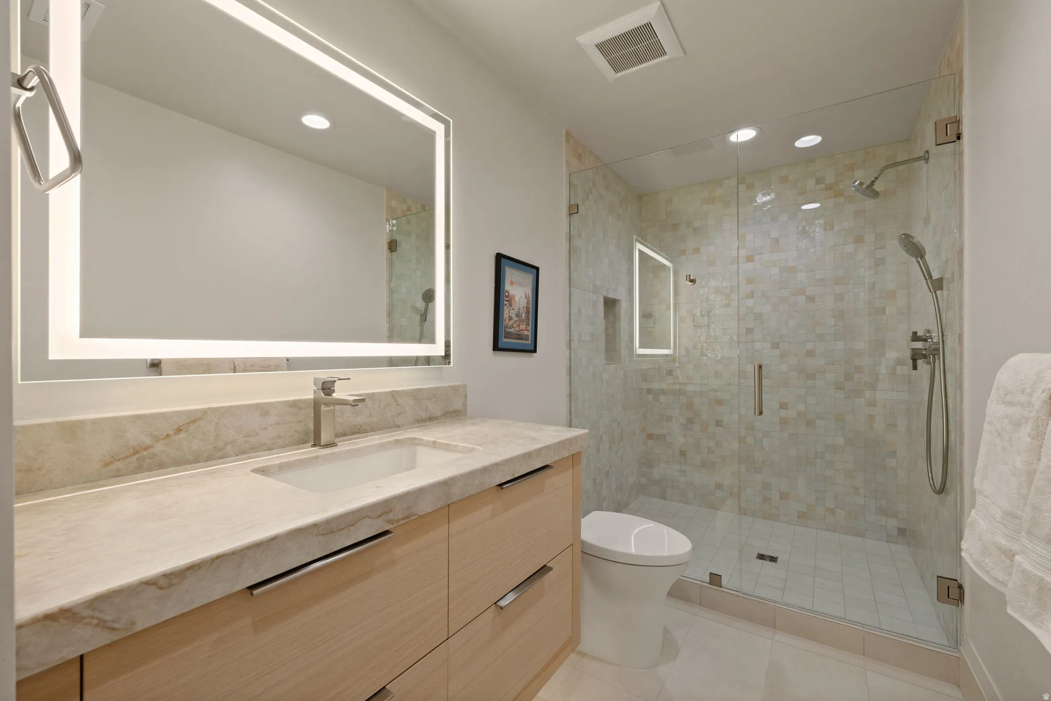 Bathroom featuring a backlit mirror, a light-toned vanity with an integrated sink, and a glass-enclosed shower with a tiled surround
