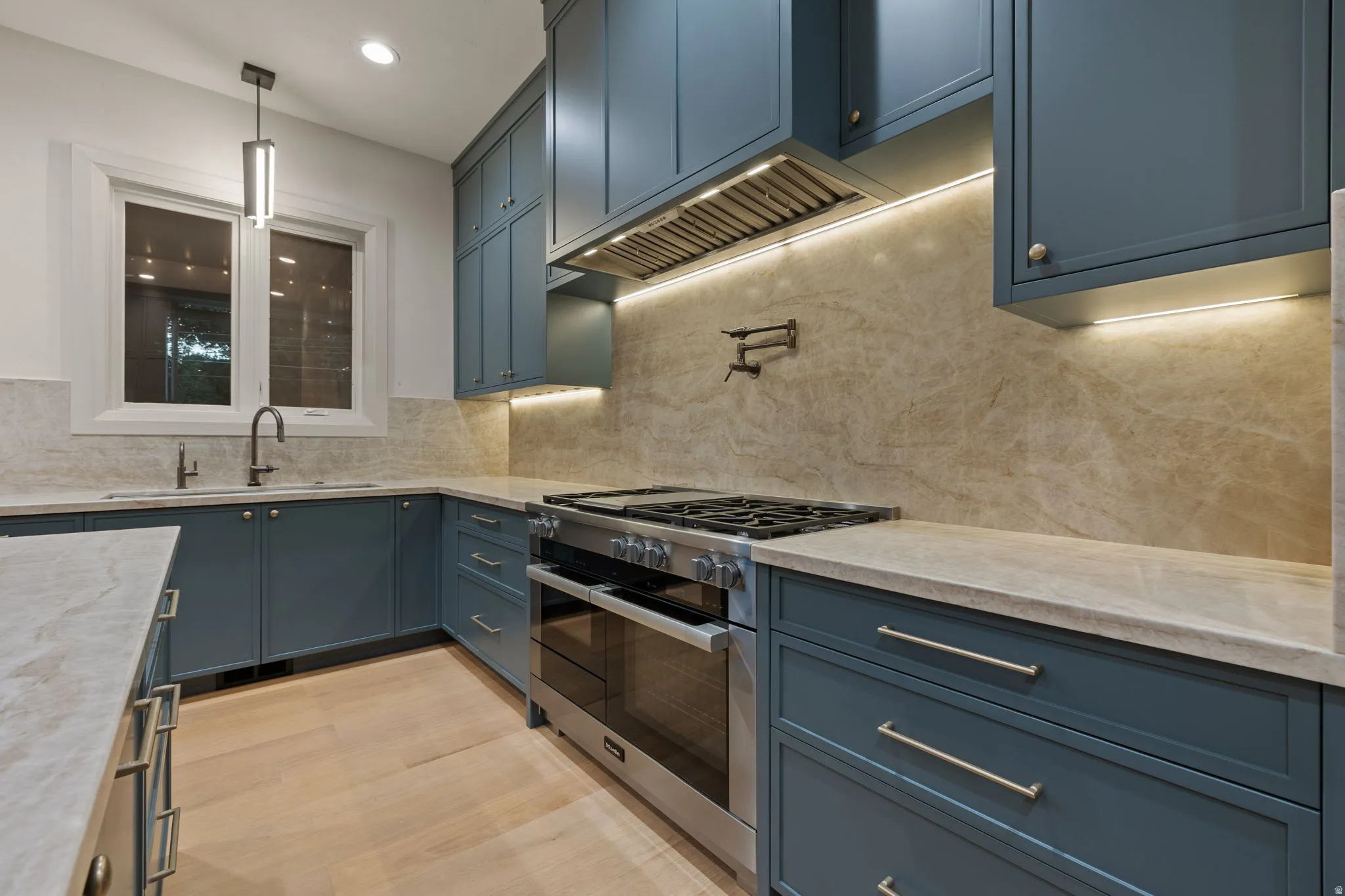 Contemporary kitchen featuring blue-gray cabinetry, light wood-finish flooring, stone countertops, and a full-height stone slab backsplash