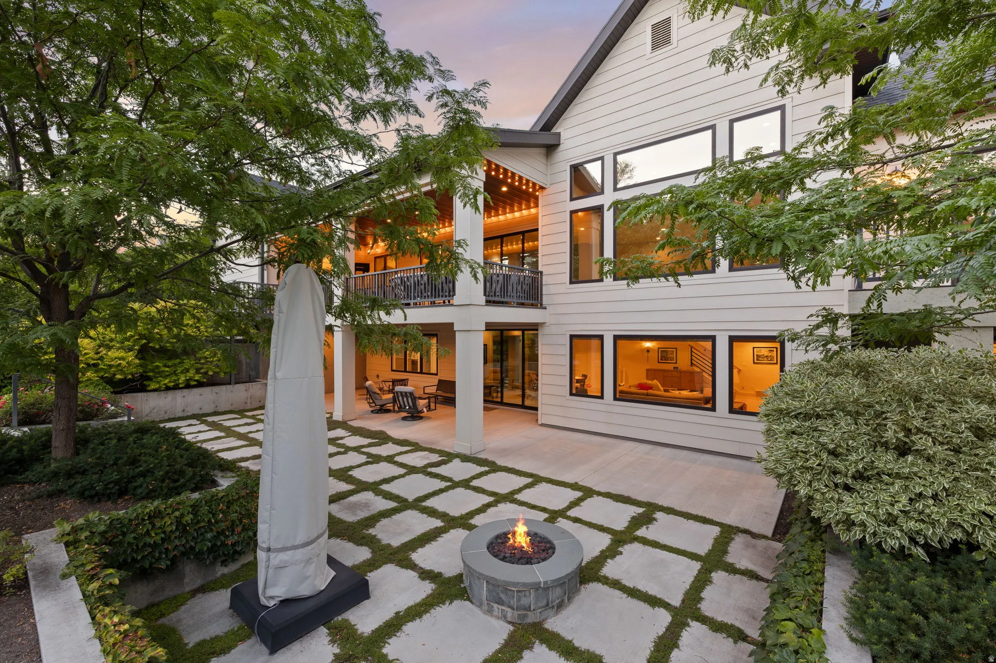 Contemporary exterior featuring an expansive concrete patio with integrated fire pit, a grid-patterned walkway with moss accents, and a covered lower-level outdoor seating area
