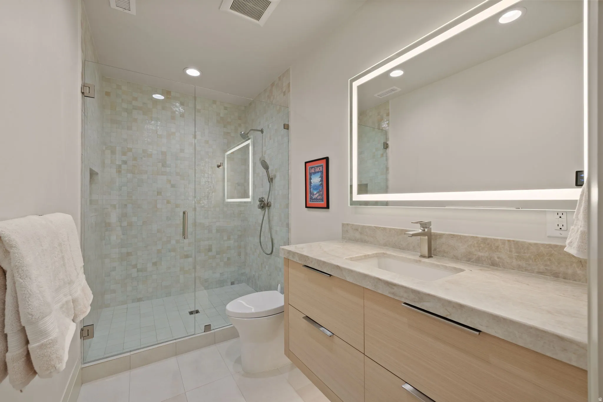 Contemporary bathroom featuring a frameless glass shower enclosure with mosaic tile, a modern vanity with light wood-finish cabinetry, a stone countertop with an integrated sink, and an illuminated mirror