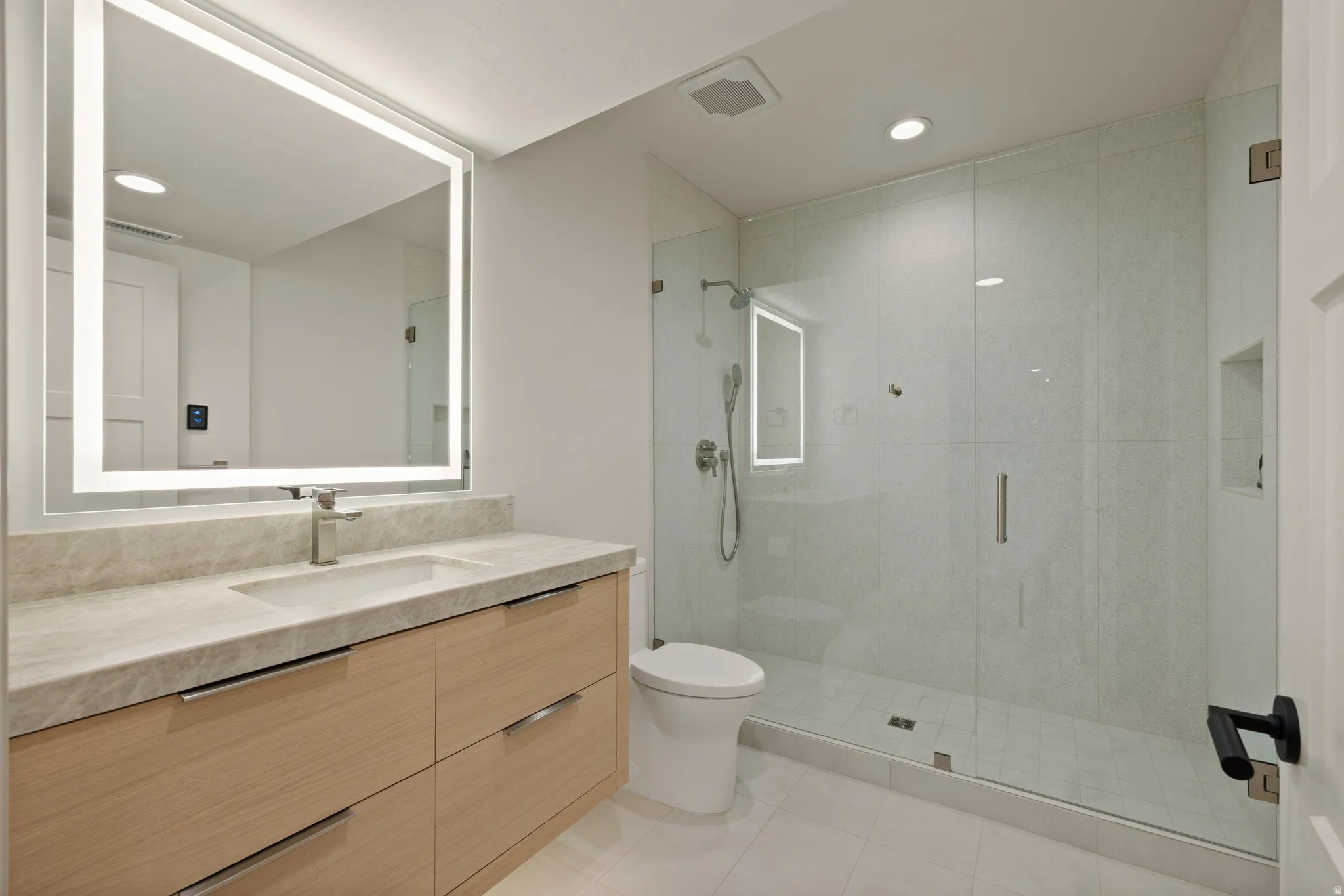 Modern bathroom featuring a wood-finish vanity with a stone countertop, an illuminated mirror, and a glass-enclosed shower