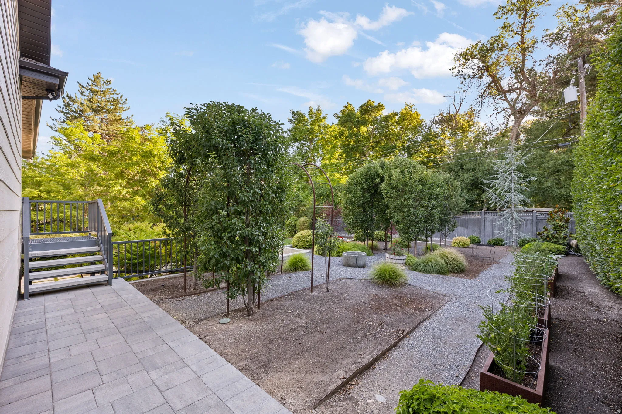 Expansive outdoor space featuring a paved patio, gravel pathways, mature trees, and raised garden beds