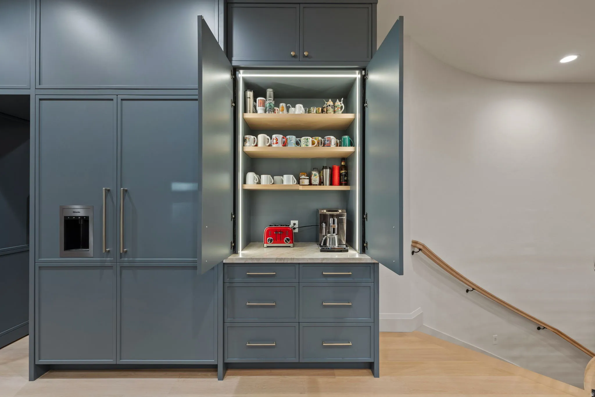 Integrated cabinetry with a built-in refrigerator and a dedicated beverage station featuring light wood shelving, task lighting, a stone countertop, and lower drawer storage