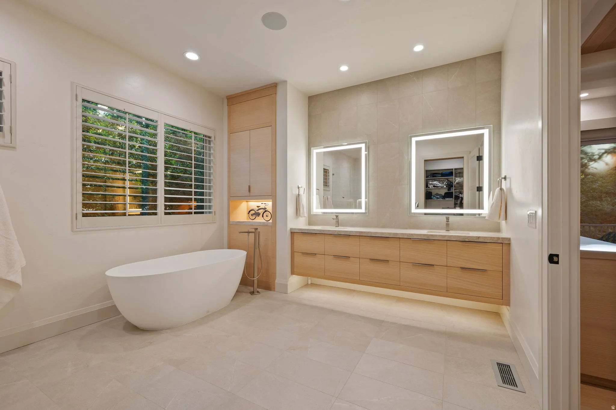 Freestanding soaking tub with floor-mounted faucet, dual illuminated vanity mirrors, double sinks, extensive wood-finish cabinetry, and large format tile flooring