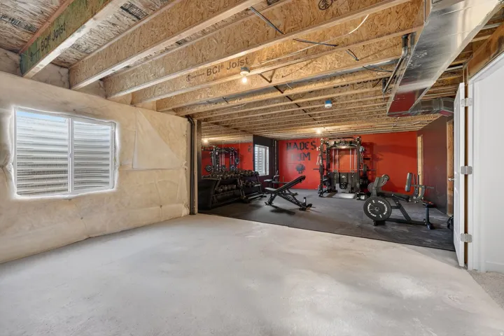Unfinished basement space with exposed ceiling joists and concrete flooring, featuring a dedicated fitness area with red accent walls and rubber flooring, and a visible egress window