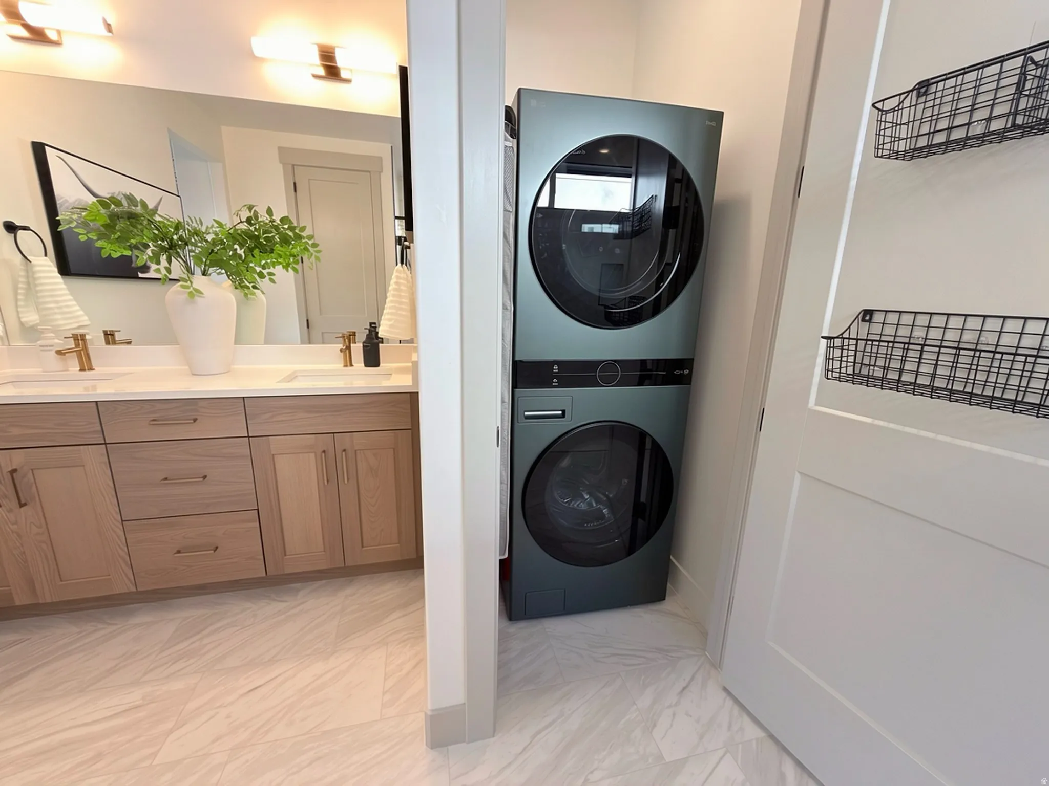 Integrated stacked washer and dryer unit, complemented by a dual vanity with light wood-finish cabinetry, white countertop, and gold-finish fixtures
