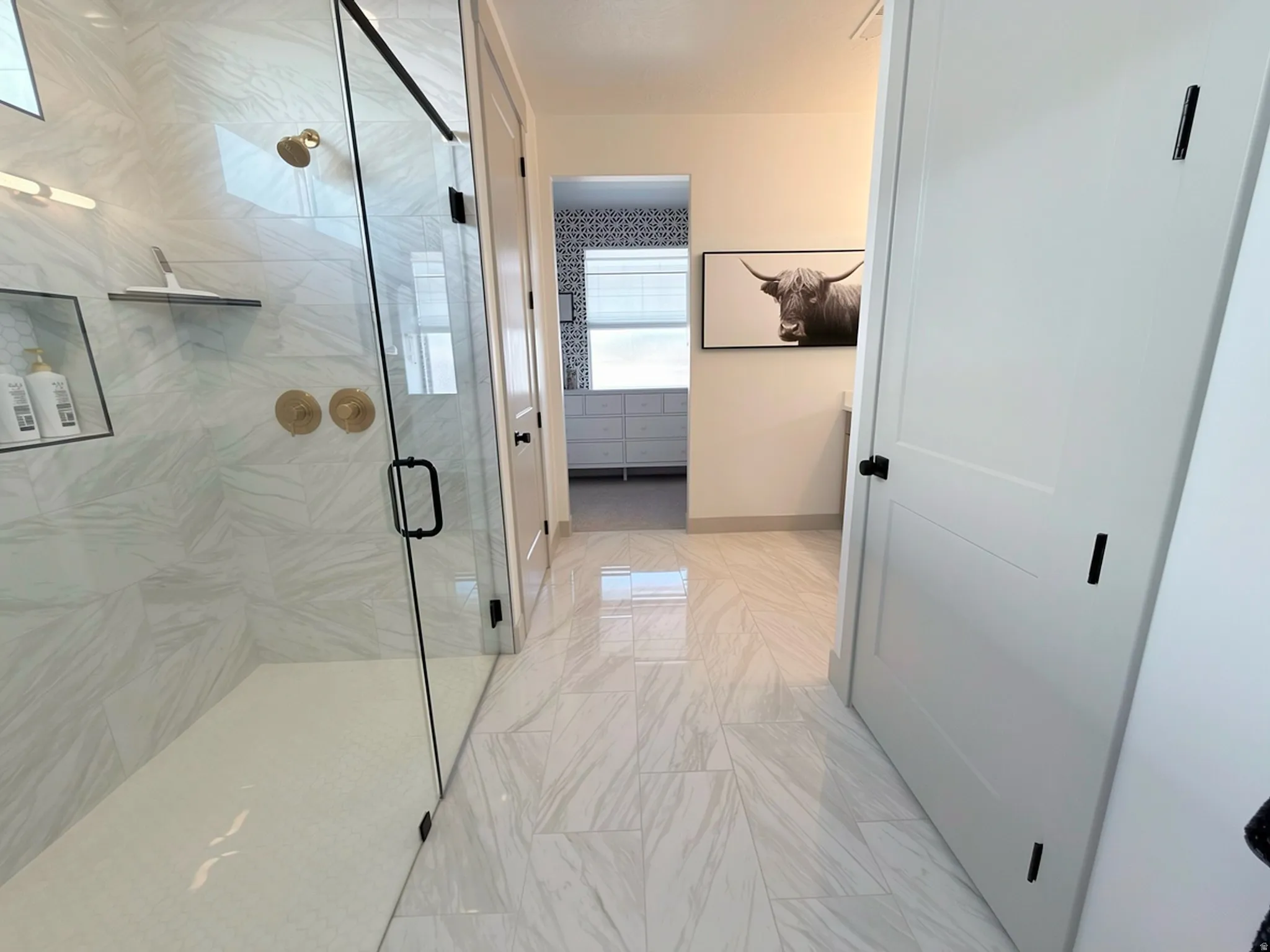 Spacious walk-in shower with clear glass enclosure and black trim, featuring large format tile with a subtle pattern and brushed gold fixtures