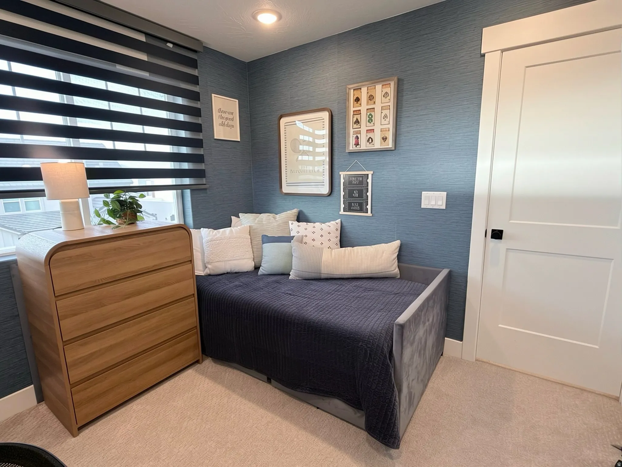 Room featuring textured blue wall covering, a window with horizontal blinds, neutral tone carpeting, and a white interior door with a black handle