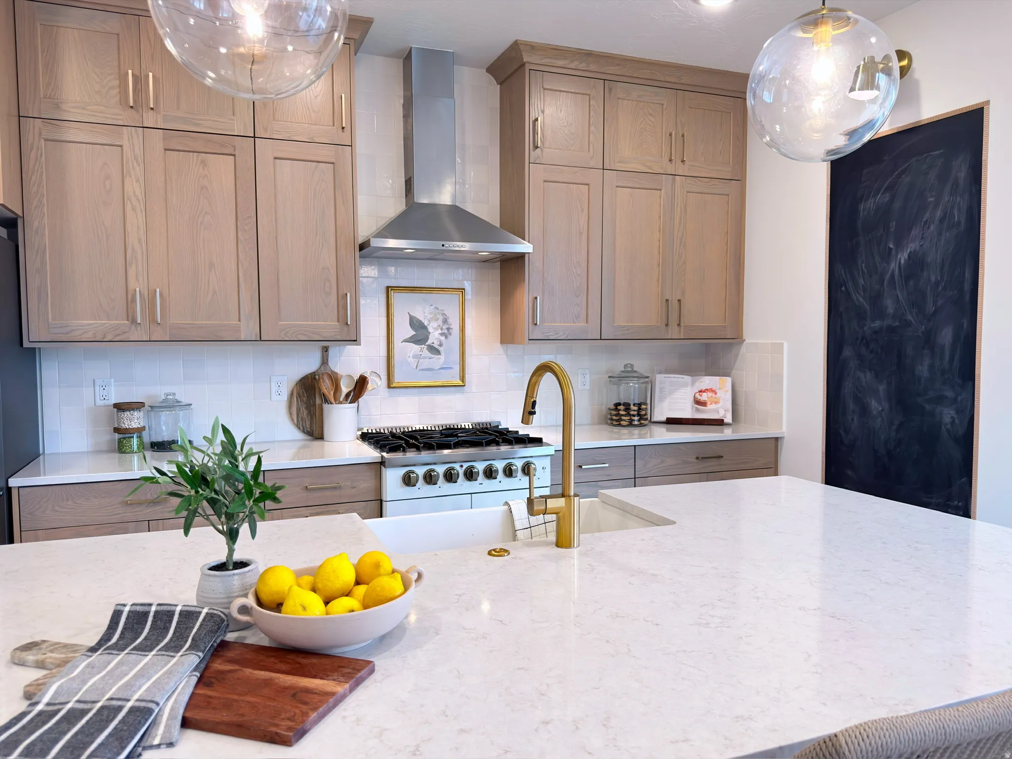 Kitchen featuring a large center island with an integrated sink, light-toned cabinetry, a stainless steel range hood, white subway tile backsplash, and a built-in chalkboard panel