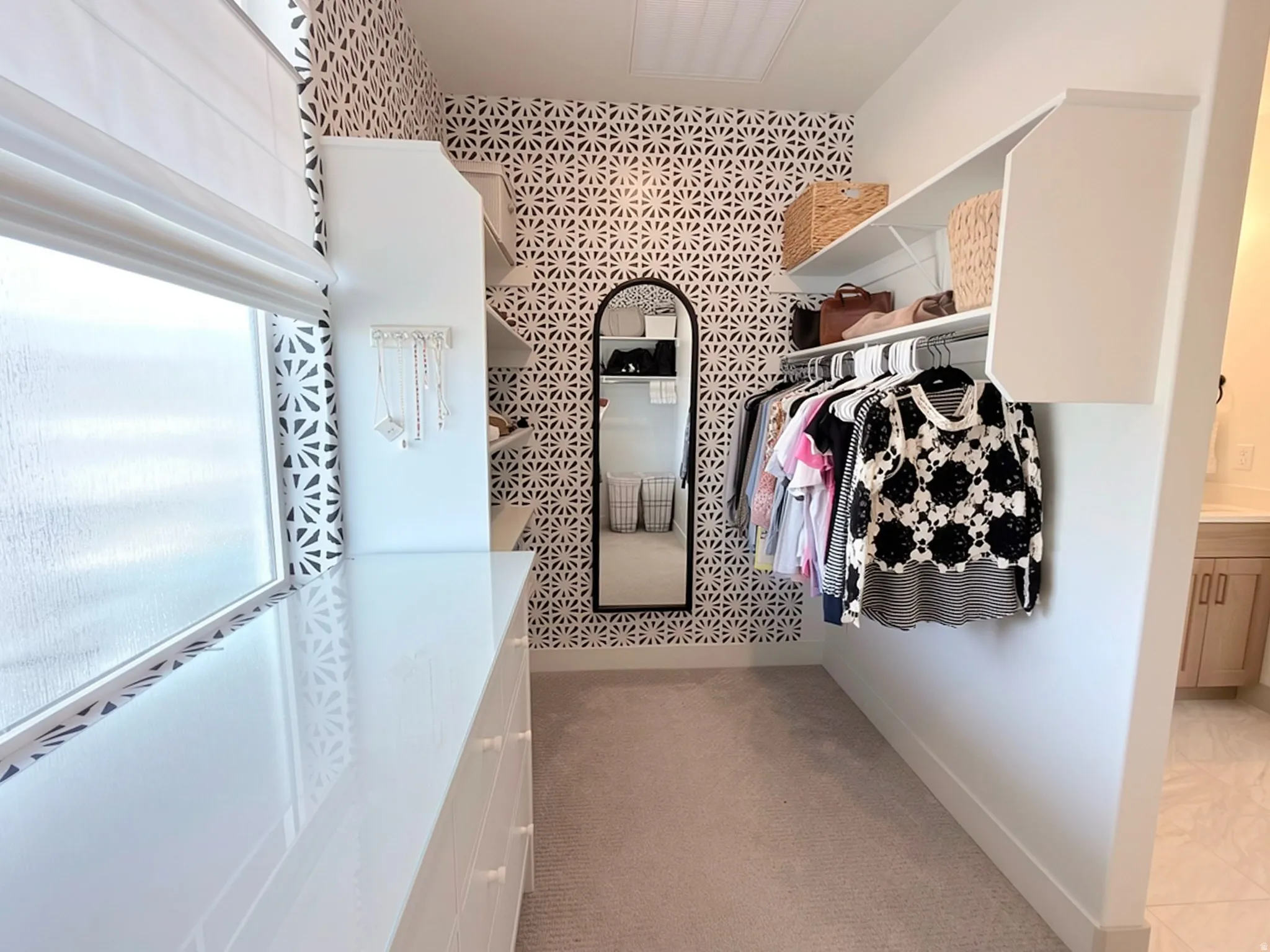 Custom closet featuring decorative wallpaper, built-in shelving, carpet flooring, a window with a roman shade, and a glossy white dresser
