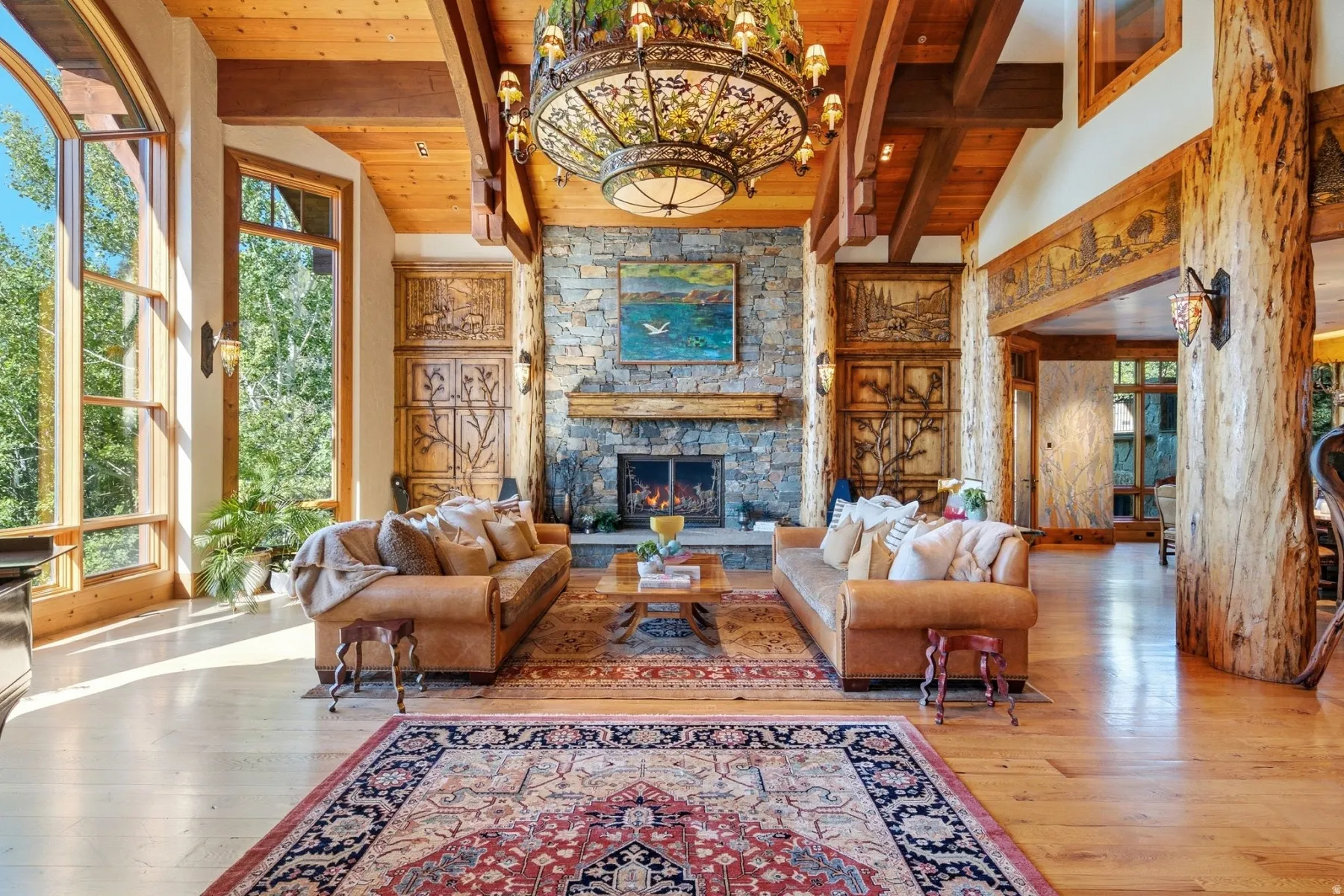Spacious living area featuring a stone fireplace with a substantial wood mantel, exposed ceiling beams, wood-finish flooring, and expansive windows