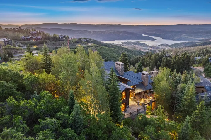 Hillside property featuring extensive tree coverage, a multi-level deck, stone chimneys, and a dark shingle roof