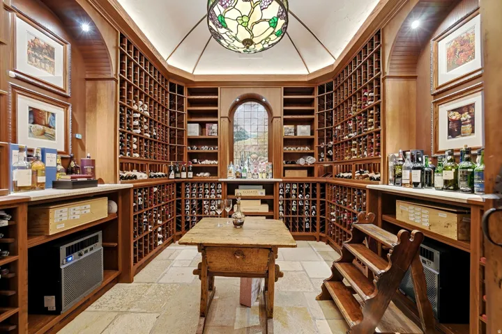 Dedicated wine cellar featuring extensive built-in wooden racking, stone tile flooring, a central wooden table, a stained-glass ceiling fixture, and a large arched window