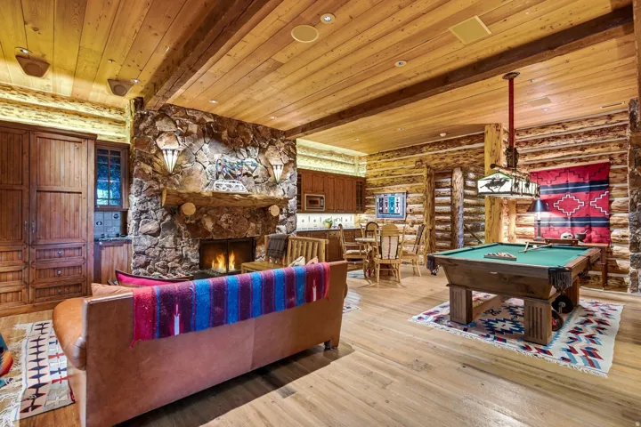 Spacious great room featuring a floor-to-ceiling stone fireplace with a substantial timber mantel, wood-finish flooring, exposed timber ceiling beams, and log-style wall construction