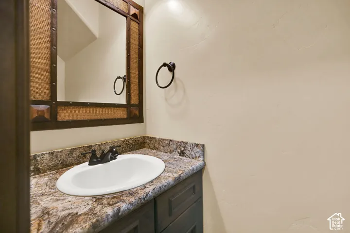 Vanity area featuring a woven and metal framed mirror, oval undermount sink, dark multi-toned countertop, and dark cabinetry with paneled doors