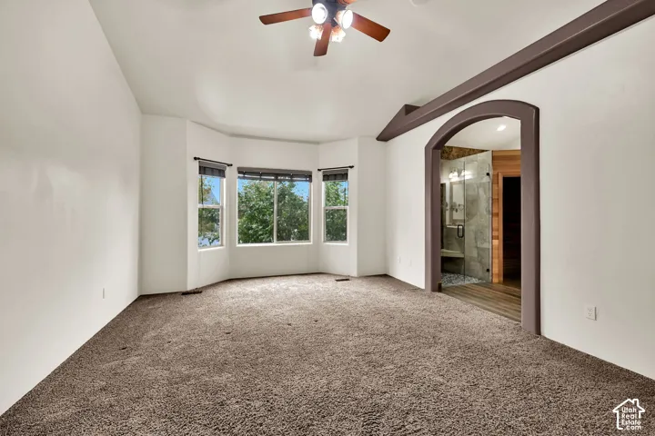 Spacious carpeted room featuring a bay window with multiple panes, a ceiling fan, and an arched doorway leading to a bathroom with a glass-enclosed shower and wood-finish flooring