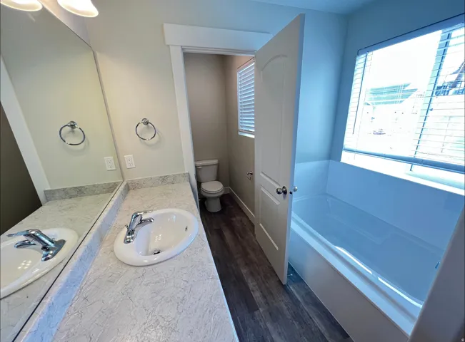 Bathroom featuring a white drop-in tub with a window, a white vanity with a composite countertop, an undermount sink, and wood-finish flooring