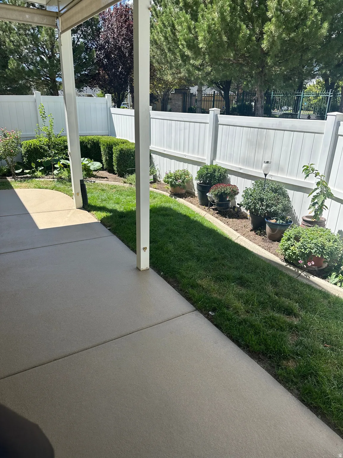 Paved patio area, manicured lawn, white privacy fencing, and mature landscaping