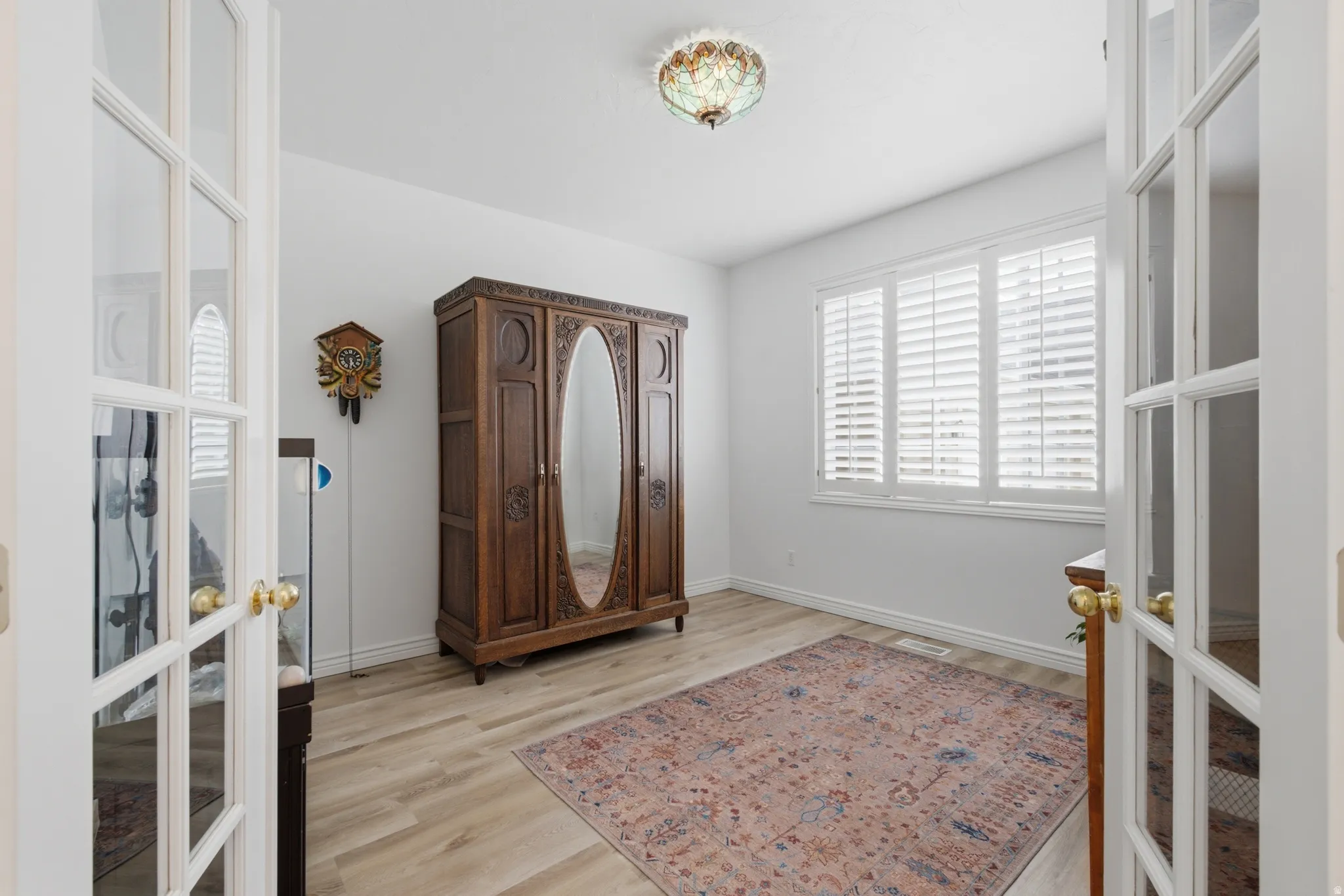 Light-filled room featuring double French doors, wood-finish flooring, a multi-pane window with plantation shutters, and a decorative ceiling light fixture