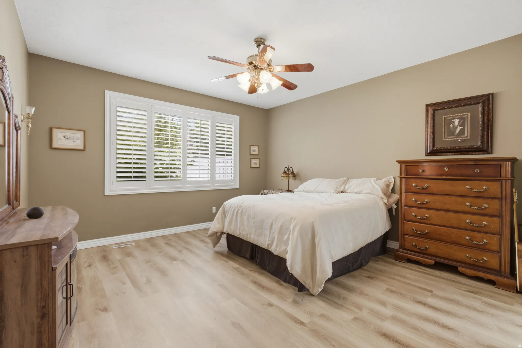 Spacious room featuring light wood-finish flooring, a multi-pane window with white plantation shutters, and a ceiling fan with integrated lighting