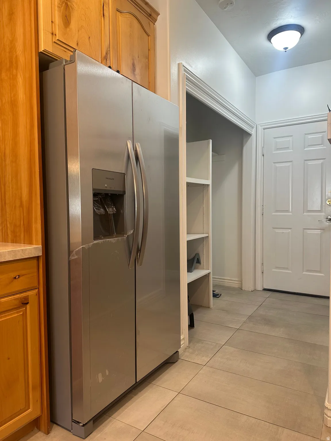 Stainless steel side-by-side refrigerator, wood cabinetry, light-toned tiled flooring, built-in shelving, and a paneled door
