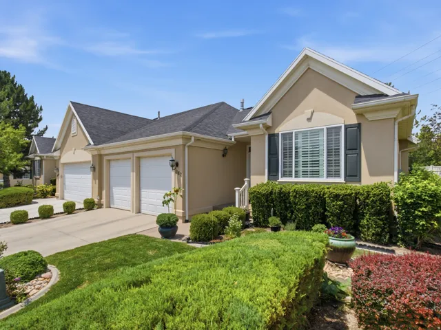 Stucco exterior with a dark shingle roof, featuring two white garage doors, a concrete driveway, and established green landscaping