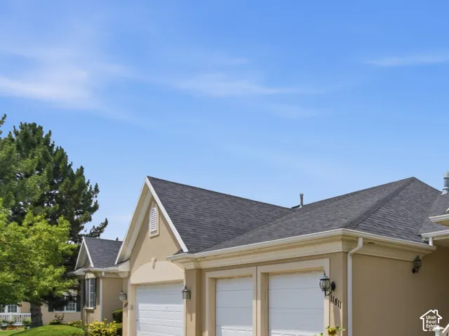 Stucco exterior with a dark shingle roof, featuring two white garage doors, a concrete driveway, and established green landscaping