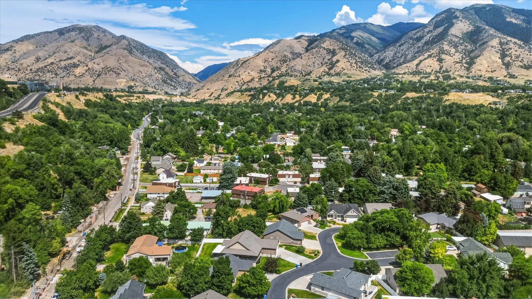 Aerial perspective of a verdant neighborhood nestled at the base of expansive mountains, featuring a mix of residential properties with mature landscaping and paved streets