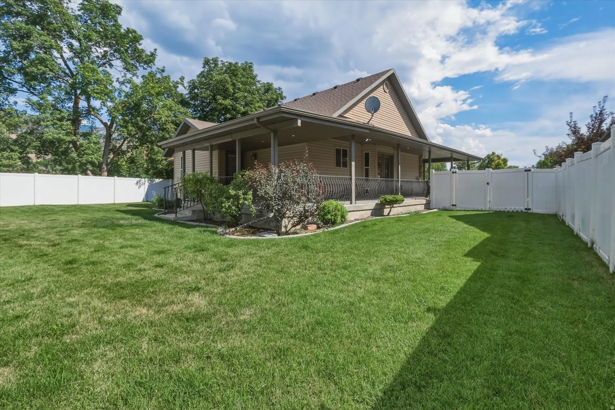 Expansive backyard featuring a green lawn, full perimeter white vinyl fencing, mature trees, and a covered patio with a railing