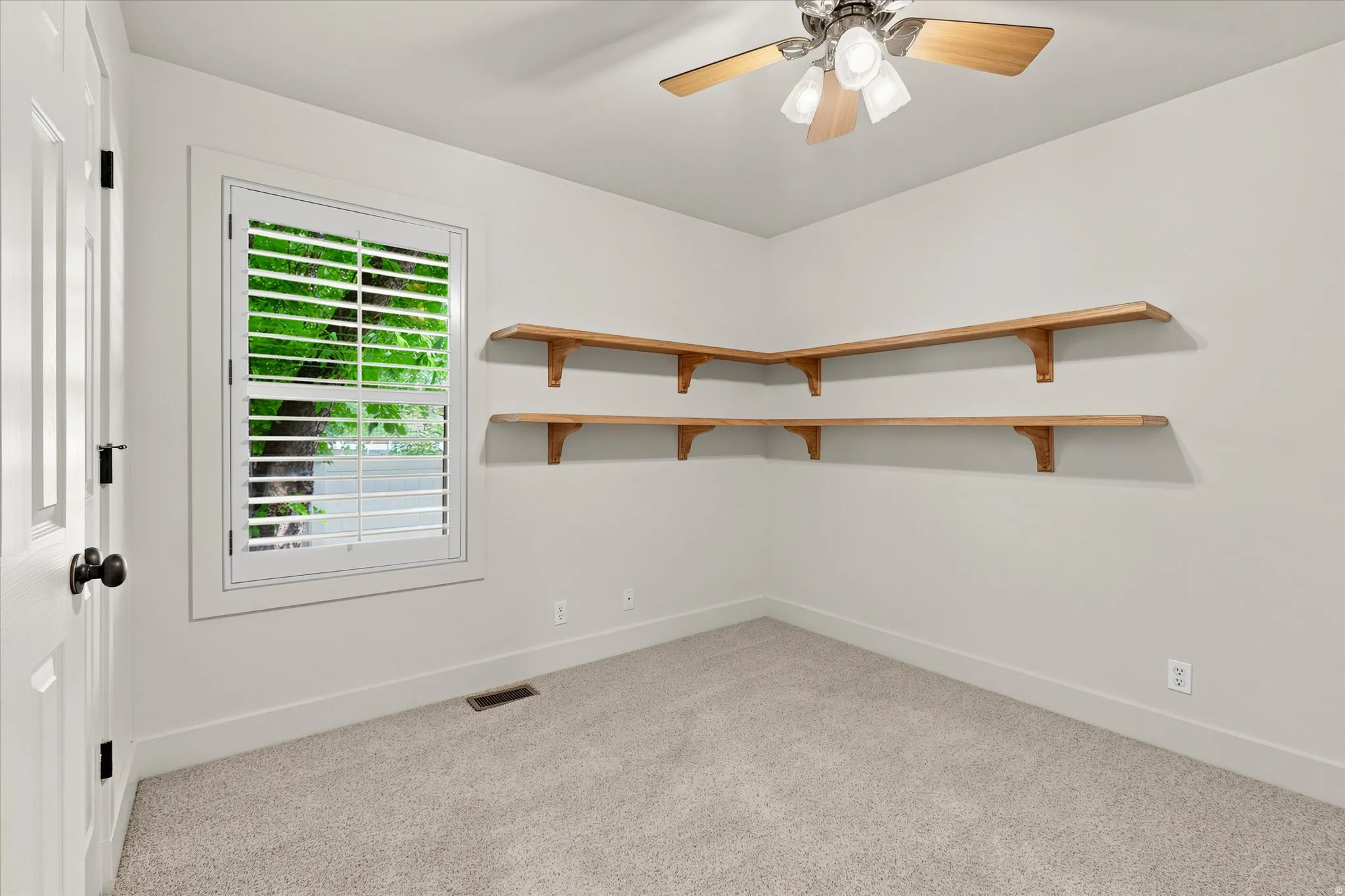 Carpeted room featuring a ceiling fan with light fixture, plantation shutters on the window, and built-in wood-finish corner shelving
