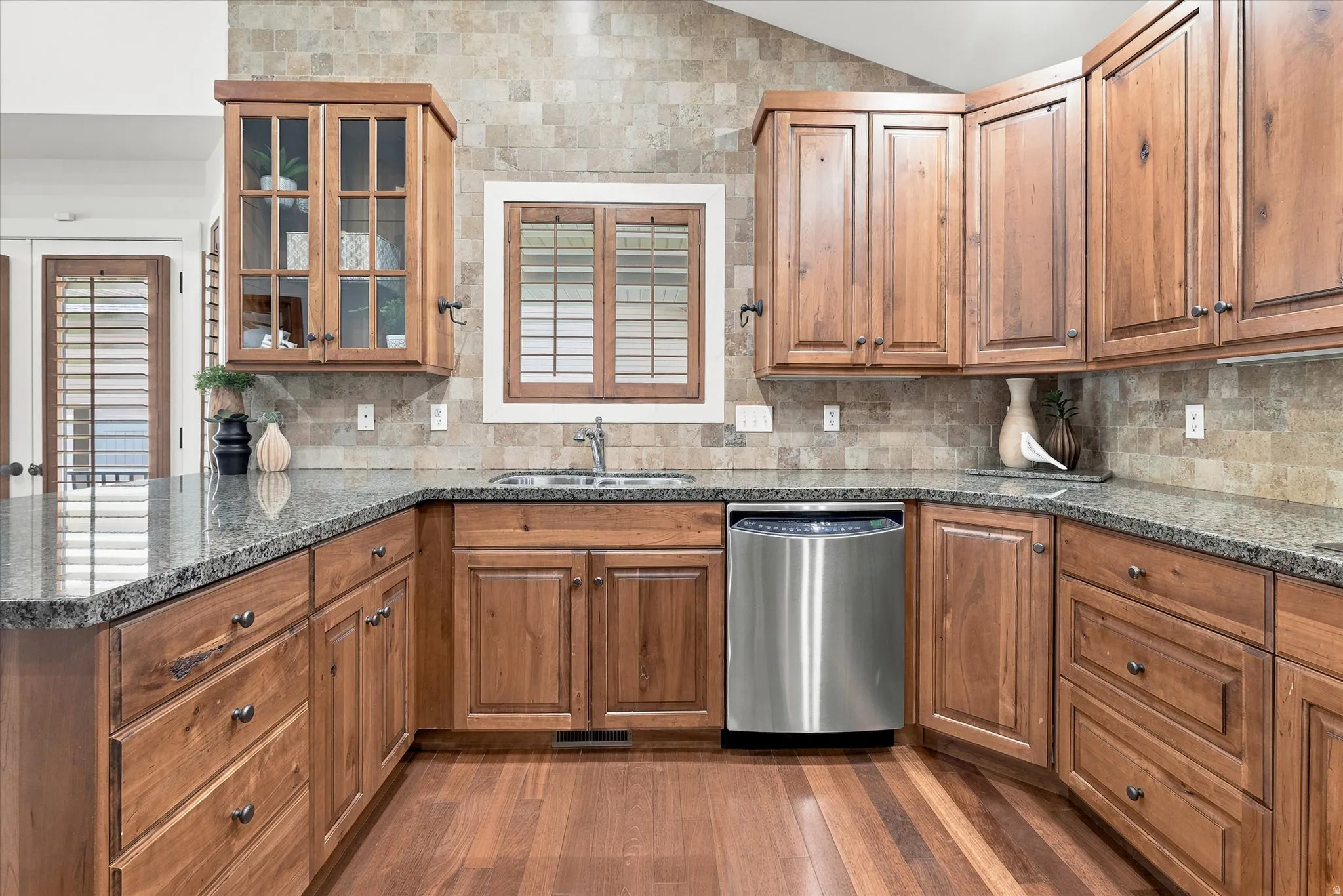 Kitchen featuring wood-finish cabinetry, stone tile backsplash, wood-finish flooring, stainless steel dishwasher, and a double basin sink