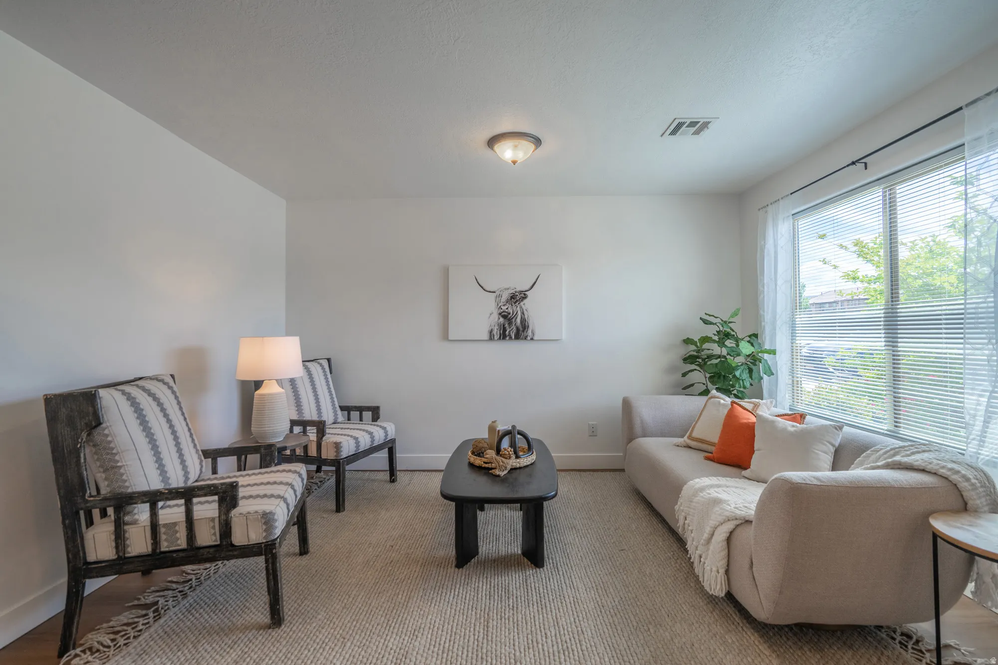 Light-filled living area featuring a broad window with blinds, neutral wall tones, recessed ceiling lighting, and wood-finish flooring partially covered by a textured area rug