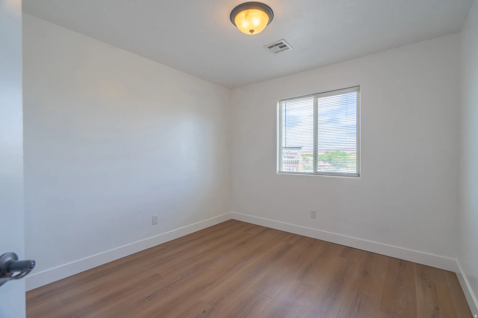 Bright interior space featuring wood-finish flooring, white walls, and a single window with blinds